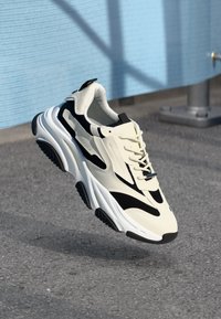 Zapatilla chunky blanca y negra con suela gruesa suspendida sobre un suelo de asfalto frente a un fondo de pared azul texturizado.