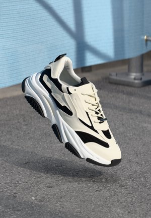 Zapatilla chunky blanca y negra con suela gruesa suspendida sobre un suelo de asfalto frente a un fondo de pared azul texturizado.