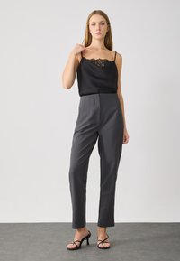 Zwarte satijnen camisole met kant, gecombineerd met hooggerimpelde grijze broek met plooien, aangevuld met zwarte hoge sandalen.