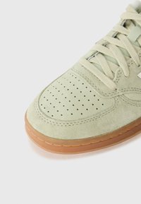 Baskets en daim vert clair avec un bout perforé, semelle beige et lacets beiges. Forme arrondie avec couture visible et détail en métal argenté.