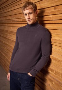 Dunkellila Rollkragenpullover mit gerippten Bündchen und Saum, kombiniert mit dunklen Jeans. Dezentes Logo-Detail auf der Brust. Strukturierter Holz-Hintergrund.