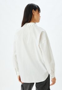 Camicia bianca a maniche lunghe con colletto button-down e borchie argentate sul colletto; texture liscia; presenta un orlo arrotondato e polsini con bottoni.