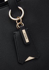 Sort faux læder håndtaske med detaljer såsom en guldfarvet metalring og et rektangulært mærke præget med "GUESS." Struktureret overflade med et elegant design.