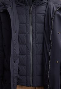 Dunkelblaues gepolstertes Jacket mit einem zentralen Reißverschluss, teilweise offener Außenschale mit Druckknöpfen und sichtbaren Nähten.
