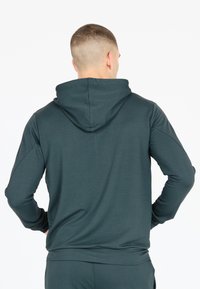 Mörkgrön huvtröja med mjuk textur, som har en känguruficka, åtsittande ärmslut och en avslappnad passform. Bakifrån betonas huvan.