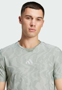 Ljusgrön sportt-shirt med ett strukturerat mönster av vågiga linjer och ränder, med en liten vit logotyp på bröstet. Korta ärmar.