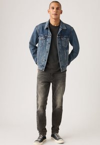 Τζιν μπουφάν μεσαίου ξεβγάλματος σε μπλε χρώμα με κλασικό κόψιμο, συνδυασμένο με μαύρο t-shirt, γκρι τζιν και μαύρα αθλητικά παπούτσια με λευκές σόλες.