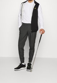 Man i vit långärmad skjorta, svart ärmlös väst, gråbyxor och svarta golfsandaler, som håller i en golfklubba och står mot en vit vägg.