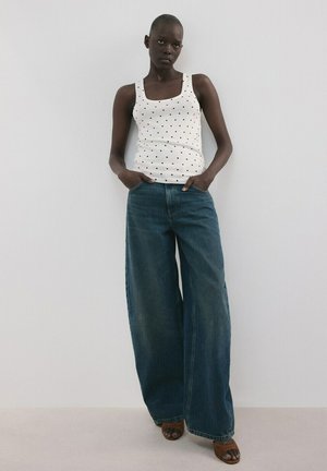 Kobieta stojąca przy białej ścianie, ubrana w biały tank top w czarne kropki, szerokie niebieskie jeansy oraz brązowe sandały z odkrytymi palcami.