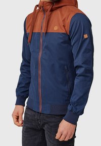 Homme portant une veste à capuche zippée bleu et rouille avec cordons et poignets côtelés, associée à un jean foncé.
