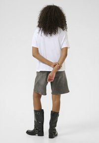 T-shirt blanc à manches courtes, shorts en denim gris, et bottes noires jusqu'aux genoux avec des accents de boucle ; cheveux bouclés, mains jointes derrière.