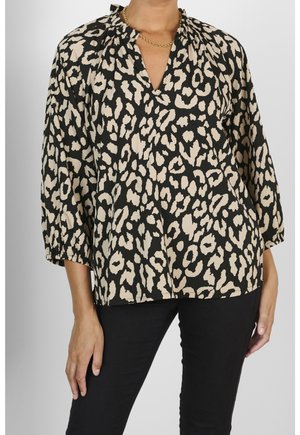 Femme portant un chemisier à imprimé léopard noir et beige avec des manches trois-quarts et un collier chaîne en or, assorti d'un pantalon noir.