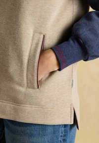 Ruka zasunutá v béžovej mikine s nápletom námořníckej farby, na ktorom sú dve červené pruhy, v kombinácii s modrými denimovými džínami.