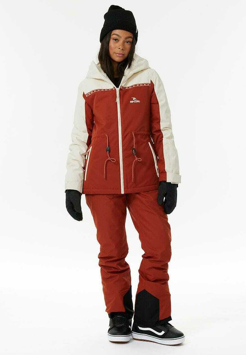 Rød og beige snow jacket med lynlås foran, justerbare snore og sorte handsker. Kombineret med røde bukser og sorte sneakers.