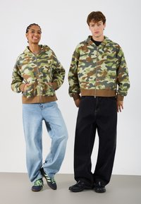 Sudaderas de camuflaje con cremalleras, puños acanalados marrones, combinadas con pantalones vaqueros holgados azules y pantalones negros. Las zapatillas presentan cordones verdes.