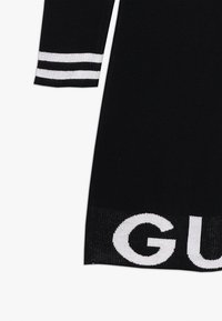 Mustan neulemekon, jossa on valkoiset raidat hihoissa ja rohkea valkoinen logo helmassa. Sujuva pinta ja vartalonmyötäinen leikkaus.