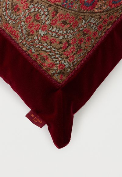 Coin d'un coussin décoratif avec une bordure en velours rouge et un centre en tapisserie florale complexe dorée, rouge et bleue sur un fond blanc.