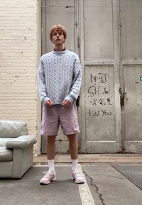 Hellblauer Kabelstrickpullover mit rundem Halsausschnitt, kombiniert mit rosa Shorts. Weiße Socken und rosa Sneakers runden das Outfit ab. Neutraler Hintergrund.