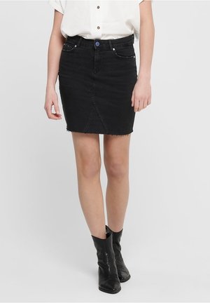 Kvinde iført en sort denim mininederdel med frynset kant, hvid kortærmet skjorte med knapper og sorte ankelstøvler, stående på hvid baggrund.