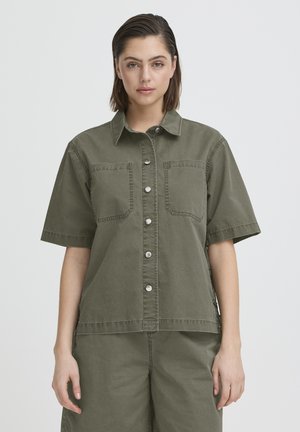 Vrouw met steil kort bruin haar, gekleed in een olijfgroen overhemd met korte mouwen en bijpassende broek, staand tegen een effen achtergrond.