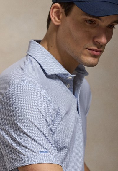 Άνδρας που φοράει ανοιχτό μπλε πουκάμισο πόλο με κοντά μανίκια και σχέδιο, και μπλε σκούφο, κοιτάζοντας προς τα κάτω με ουδέτερη έκφραση.