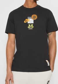 Svart Nike t-shirt med grafik av en solros, gröna blad, jord och en orange Nike-logotyp på bröstet, bärs med vita byxor.