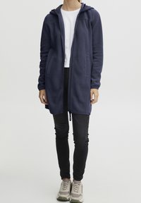 Abrigo largo de forro polar azul marino con capucha, cremallera frontal y dos bolsillos laterales, llevado sobre una camisa blanca y pantalones negros, combinado con zapatillas deportivas grises.