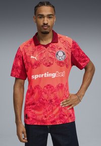 Jersey atlético rojo con un patrón floral, de mangas cortas y con un collar. Incluye el logotipo de Puma y el texto "sportingbet" en la parte frontal.