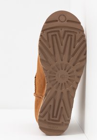 Semelle de chaussure en daim marron avec un motif de bande de roulement en zigzag profond et un design circulaire autour du logo UGG au centre.