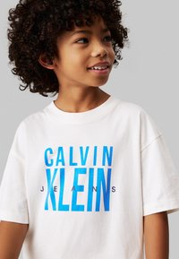 T-shirt bianco in cotone con maniche corte, con una grafica blu audace che riporta la scritta "CALVIN KLEIN JEANS" in due tipi di carattere.