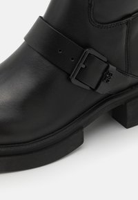 Černá kožená bota s plochou podrážkou, širokým kotníkem a bočním páskem s obdélníkovou sponou. Hladký povrch bez viditelných vzorů.