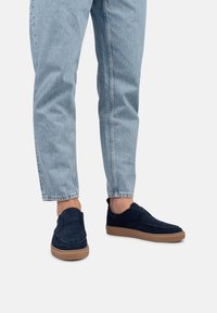 Zapatos slip-on de ante azul marino con suela de goma, que presentan detalles de costura y una textura suave. Combinados con jeans de mezclilla de color azul claro.