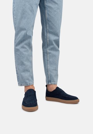 Mocassins en daim bleu marine avec une semelle en caoutchouc, présentant des détails cousus et une texture lisse. Portés avec un jean en denim bleu clair.