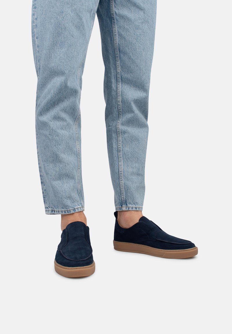 Zapatos slip-on de ante azul marino con suela de goma, que presentan detalles de costura y una textura suave. Combinados con jeans de mezclilla de color azul claro.