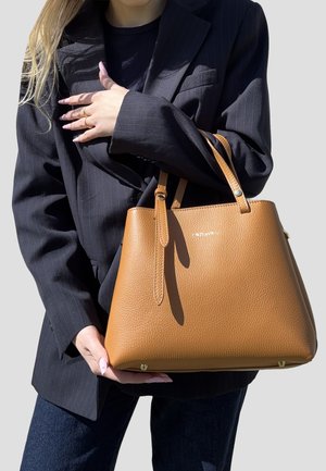 Persona con blazer nero e jeans tiene una grande borsa in pelle marrone chiaro con due manici su uno sfondo semplice.