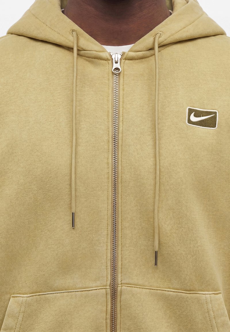 Mann trägt beigefarbene Kapuzenjacke mit Reißverschluss und Kordeln sowie kleinem Nike-Logo auf der linken Brustseite, sichtbar vom Hals bis zur Taille.