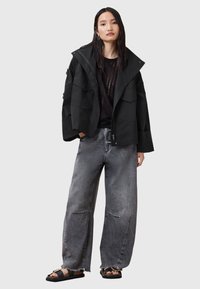Giacca impermeabile nera con grandi tasche e un ampio colletto, abbinata a jeans grigi larghi con orlo sfrangiato e vestibilità comoda. Sandali inclusi.