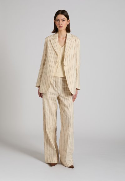 Frau trägt cremefarbenen Nadelstreifen-Übergröße-Blazer und weit geschnittene Hose mit cremefarbenem Strick-V-Ausschnitt-Oberteil, steht vor schlichtem grauem Hintergrund.
