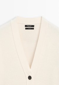 Crème-kleurige cardigan gemaakt van 100% wol, met een V-hals, geribbelde textuur en een enkele zwarte knoopsluiting. Merkenlabel binnenin.