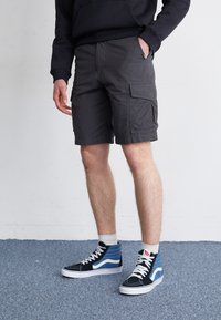 Pantalones cortos de carga grises con bolsillos laterales, textura de la tela visible. Combinados con zapatillas altas en azul marino y azul, con cordones y suela blanca.