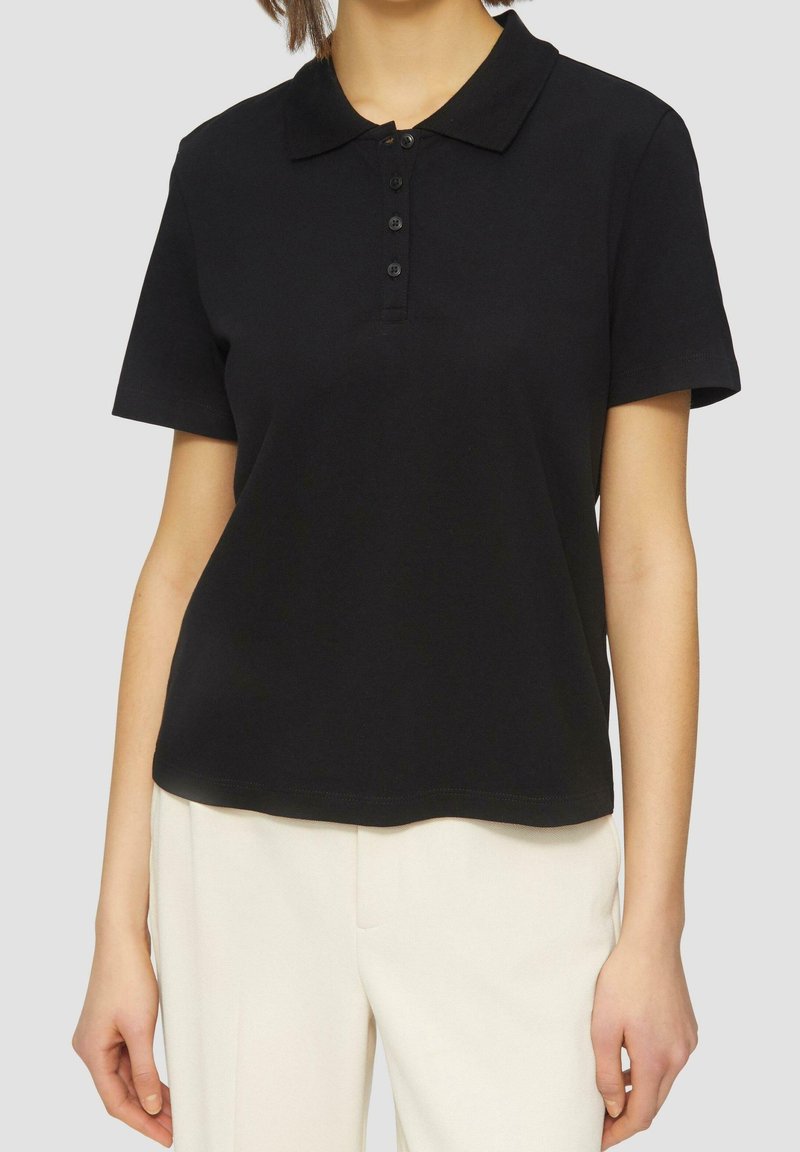 Personne portant un polo noir à manches courtes avec quatre boutons, associé à un pantalon blanc taille haute, visible du cou jusqu'aux hanches.
