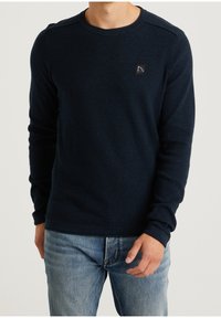 Granatowy sweter z dzianiny z okrągłym dekoltem, długimi rękawami, teksturowanym wzorem i małą naszywką z logo na klatce piersiowej.