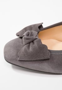 Chaussure plate en suède gris avec un bout arrondi, mettant en avant un détail de nœud prononcé à l'avant et une semelle intérieure lisse de couleur claire.