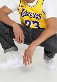 Maglia da basket gialla con la scritta "Lakers" e il numero "23", abbinata a jeans neri in denim e sneakers bianche. Cuffie dei pantaloni arrotolate.