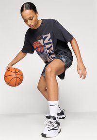 T-shirt Nike supradimensionat, de culoare neagră, cu grafică de baschet, pantaloni scurți albaștri cu detalii din plasă, șosete albe Nike și adidași negri cu alb. Ținând o minge de baschet.