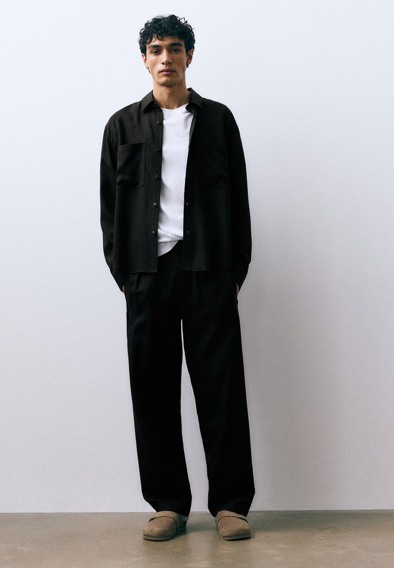 Jeune homme aux cheveux bouclés portant une chemise noire ouverte sur un t-shirt blanc, un pantalon noir et des chaussures beige à enfiler, debout contre un mur blanc.