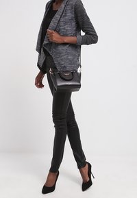 Svart handväska med en strukturerad design, med roséguld detaljer. Modellen bär en grå strukturerad jacka, figursydda svarta byxor och svarta klackskor.