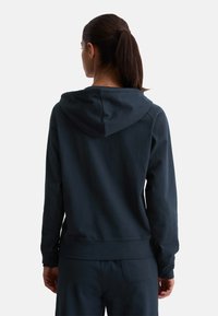 Mujer con el cabello oscuro en una coleta, vistiendo una sudadera oscura sencilla de color azul marino y pantalones a juego, mostrada de espaldas contra un fondo blanco.