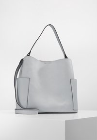 Sac seau en cuir gris clair avec une finition texturée, doté de deux poches latérales et d'une bandoulière lisse, design minimaliste sans ornements.