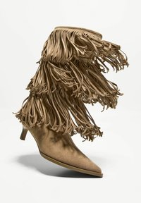 Brązowy zamszowy botek za kostkę z frędzlami, zaostrzonym noskiem i krótkim obcasem typu słupek. Charakteryzuje się lekką fakturą i płynnymi akcentami frędzli.